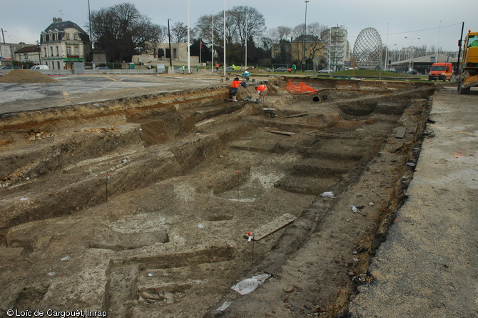 Au premier plan, le long de la berne, on distingue une voie médiévale. En contrebas s'alignent différentes structures gallo-romaines du Ier s. de notre ère. Place de la République, Reims (Marne), 2007-2008.  Les constructions antiques bordaient le cardo maximus (la principale voie nord-sud) de la cité. A l'époque médiévale, le creusement d'un fossé et son comblement suivi de la mise en place d'une voie en partie empierrée ont perturbé les niveaux archéologiques antiques. 