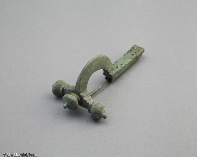 Fibule cruciforme en bronze, longueur 7 cm, fin du IVe-début du Ve siècle, mis au jour dans le sanctuaire du site de la ZAC du Clos de la Fontaine de l'Étuvée à Orléans (Loiret). Ces fibules sont caractéristiques du vêtement des fonctionnaires civils et militaires. 