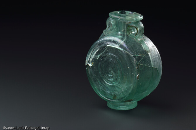 Flacon en verre souffl&eacute; dans un moule, 170-250 de notre &egrave;re, mis au jour lors de la fouille de la Zac Pasteur &agrave; Besan&ccedil;on (Doubs) entre 2010 et 2011. Le fond est orn&eacute; de l'inscription COM.F correspondant peut-&ecirc;tre &agrave; l'abr&eacute;viation d'un nom de potier. 