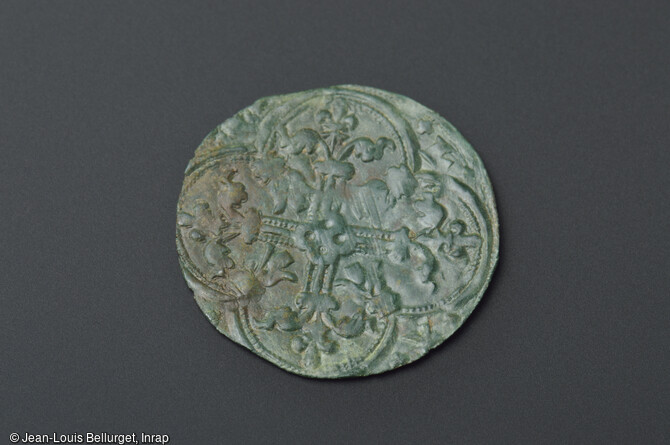 Jeton de commerce en cuivre, 1326-1400, d&eacute;couvert lors de la fouille de la Zac Pasteur &agrave; Besan&ccedil;on (Doubs) entre 2010 et 2011. Cette face porte une croix dont chaque branche est termin&eacute;e par une fleur de lys. L'autre face, montre un roi couronn&eacute; assis sur une chaise gothique. 