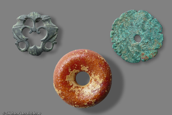 &Agrave; gauche, applique de harnais ajour&eacute;e de type  trompette muster  repr&eacute;sentant des dauphins affront&eacute;s (3e si&egrave;cle de notre &egrave;re). &Agrave; droite, rondelle dent&eacute;e retrouv&eacute;e dans un vase avec trois fibules et interpr&eacute;t&eacute;e comme applique vestimentaire. En dessous, perle en ambre issue d'une s&eacute;pulture, exceptionnelle par ses dimensions (3,8 cm de diam&egrave;tre). D&eacute;couvertes lors de la fouille &agrave; Saint-Sauveur (Somme) en 2015.