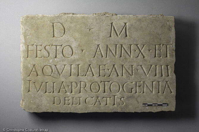 Plaque en marbre qui porte l'&eacute;pitaphe &laquo; Aux Dieux M&acirc;nes. A Festus, &acirc;g&eacute; de dix ans, et &agrave; Aquila, &acirc;g&eacute; de huit ans, Iulia Protogenia, &agrave; ses ch&eacute;ris &raquo;. Elle a &eacute;t&eacute; d&eacute;couverte dans une s&eacute;pulture &agrave; Narbonne (Aude).  Une n&eacute;cropole antique (1er au 3e si&egrave;cle) a livr&eacute; des vestiges fun&eacute;raires exceptionnels tant par leur nombre (pr&egrave;s de 500 tombes) que par leur &eacute;tat de conservation.  