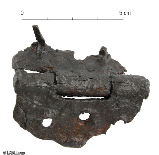 Charnière en fer découverte dans la cave antique du site de Drulingen. Elle est constituée de tôles rectangulaires fragmentaires articulées avec trois charnons. Les perforations et le rivet de fixation sont visibles. Le mobilier mis au jour dans les différents comblements de la cave permet d’envisager son aménagement en bois et d’appréhender son ameublement (restes de coffre).