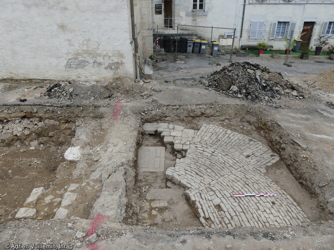 Vue des vestiges de la fontaine accolée contre le mur sud de l'église en cours de fouille à Baume-les-Dames (Doubs) édifiée entre la seconde moitié du 17e siècle et le 18e siècle. En subsiste le socle monolithique associé à un conduit d'évacuation, le massif de fondation du bassin de plan semi-ovalaire, et un sol pavé se déployant alentour.