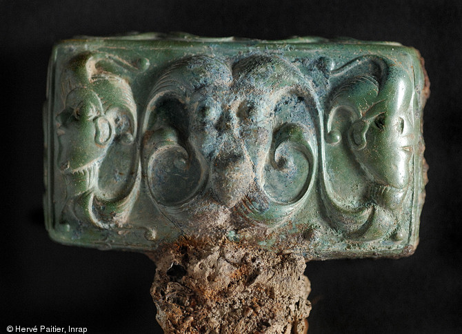 T&ecirc;te de clavette de char en bronze, d&eacute;cor&eacute;e en bas-relief d'un visage vu de face encadr&eacute; par deux profils. Cette pi&egrave;ce appartient au style &laquo;&nbsp;plastique&nbsp;&raquo; dat&eacute; de la fin de La T&egrave;ne ancienne (environ 300-250 avant notre &egrave;re).  D&eacute;couverte durant l'&eacute;t&eacute; 2006, la tombe d'Orval (Manche) est aujourd'hui un cas unique&nbsp;: la plus occidentale des &laquo;&nbsp;tombes &agrave; char&nbsp;&raquo; de la fin de La T&egrave;ne ancienne jamais trouv&eacute;e en Europe est aussi la seule de Gaule celtique &agrave; contenir des chevaux. 