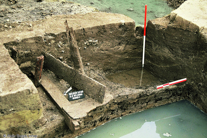 Bief bordé de deux alignements de pieux, débouchant sur un bassin par l'intermédiaire d'une vanne composée d'un assemblage de planches, pieux et madriers, milieu du VIIIe s., Saleux (Somme), 1993.  Cet aménagement est un témoin de la présence d'un moulin à eau. Ce type de moulin, mentionné par les textes médiévaux, est rarement mis en évidence par l'archéologie.  Photo publiée dans l'ouvrage Archéologie médiévale en France - Le premier Moyen Âge, Isabelle Catteddu, coll. Archéologies de la France. 