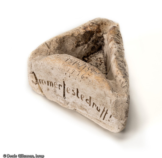 Artisanat de tranchée : cendrier triangulaire taillé dans un bloc de craie, face latérale gauche avec inscription  Immer Feste Drüff  (Allez-y ferme), formule utilisée en patois berlinois-brandebourgeois par le Konprinz Guillaume suite aux événements de Saverne en 1913. Découvert dans une latrine d'un terrain d'aviation allemand datant de la Grande Guerre à Warmeriville (Marne), 2013.