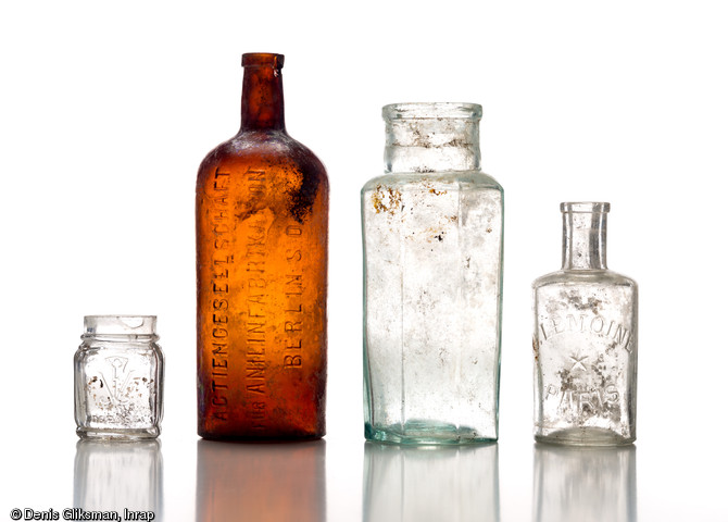 Quatre flacons en verre. De gauche à droite : petit flacon marqué  VF-Déposé , pommade ou baume indéterminé ; bouteille d'aniline avec inscription  Actiengesellschaft für Anilinfabrikation Berlin S.O.  (Société de fabrication d'aniline, Berlin) l'aniline est un composé organique aromatique de formule chimique utilisé pour la teinture des tissus ; flacon de forme octogonale sans marque ; petit flacon de fortifiant (?) sans marque. Découverts dans une latrine d'un terrain d'aviation allemand datant de la Grande Guerre à Warmeriville (Marne), 2013.