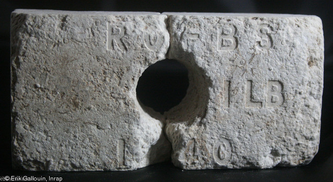 Brique en papier compress&eacute;, estampill&eacute;e et perfor&eacute;e en son centre de sorte &agrave; pouvoir &ecirc;tre empil&eacute;e sur une armature en bois, d&eacute;couverte dans un trou d'homme &agrave; Cagny (Calvados), 2013.  Produites depuis l'entre-deux-guerres jusque dans les ann&eacute;es 50, ce genre de brique a servi &agrave; la construction rapide de baraquements pendant la guerre et de maisons pour abriter les familles sans-logis dans l'imm&eacute;diat-apr&egrave;s-guerre.  Ces mat&eacute;riaux produits en s&eacute;rie ont &eacute;t&eacute; import&eacute;s des Etats-Unis et du Canada. 